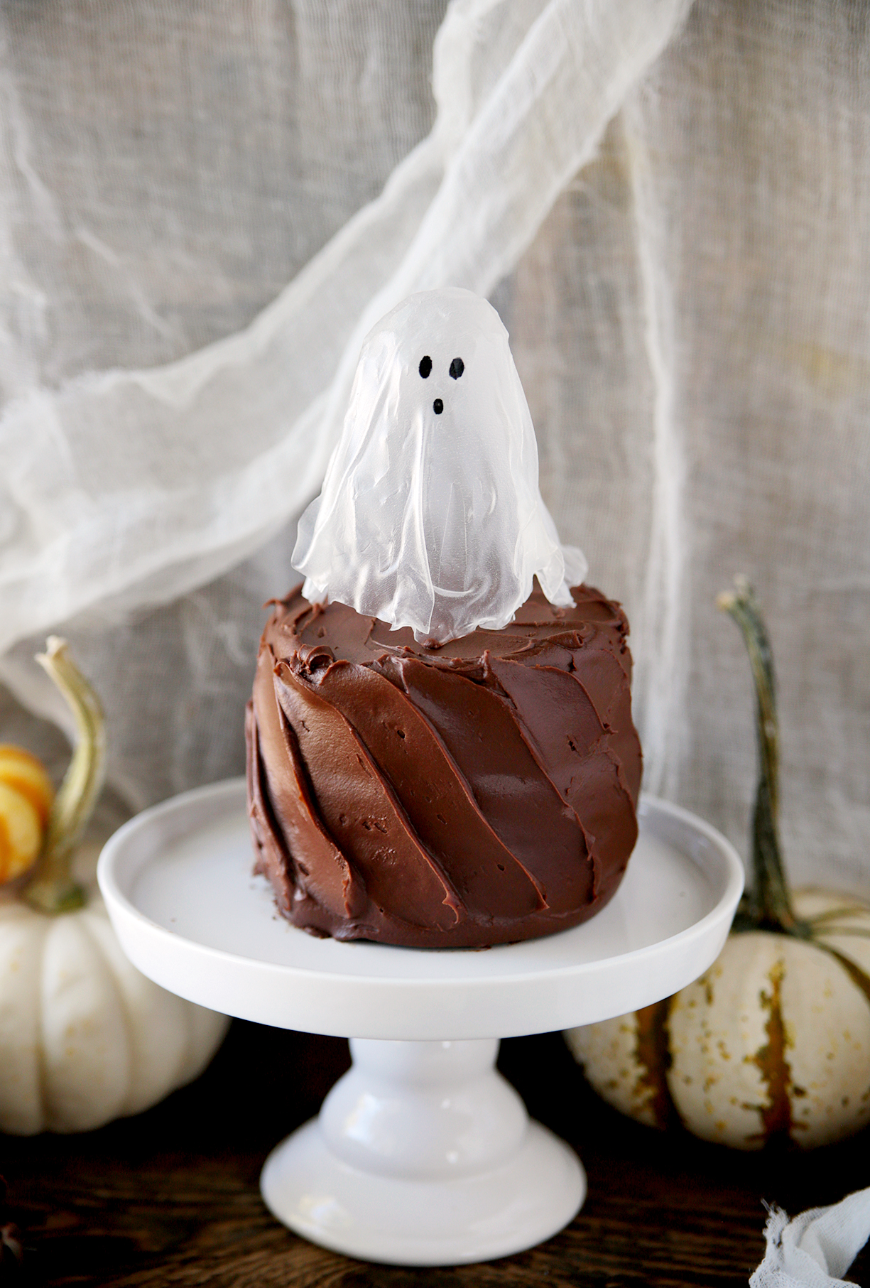 rice paper ghost on small chocolate cake on white cake stand with mini pumpkins sitting behind cake stand